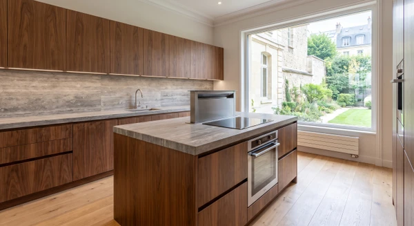 Photographie d'une cuisine sur mesure moderne en noyer américain veiné, façades sans poignées apparentes avec détails de quincaillerie Blum intégrée. Plan de travail en pierre naturelle grise type travertin, crédence assortie. Îlot central avec plaque induction encastrée, four intégré en inox, hotte discrète intégrée. Parquet massif chêne clair, éclairage LED sous-meuble, baie vitrée apportant lumière naturelle zénithale. Mobilier et agencement montrant le savoir-faire artisanal et la qualité des matériaux durables.