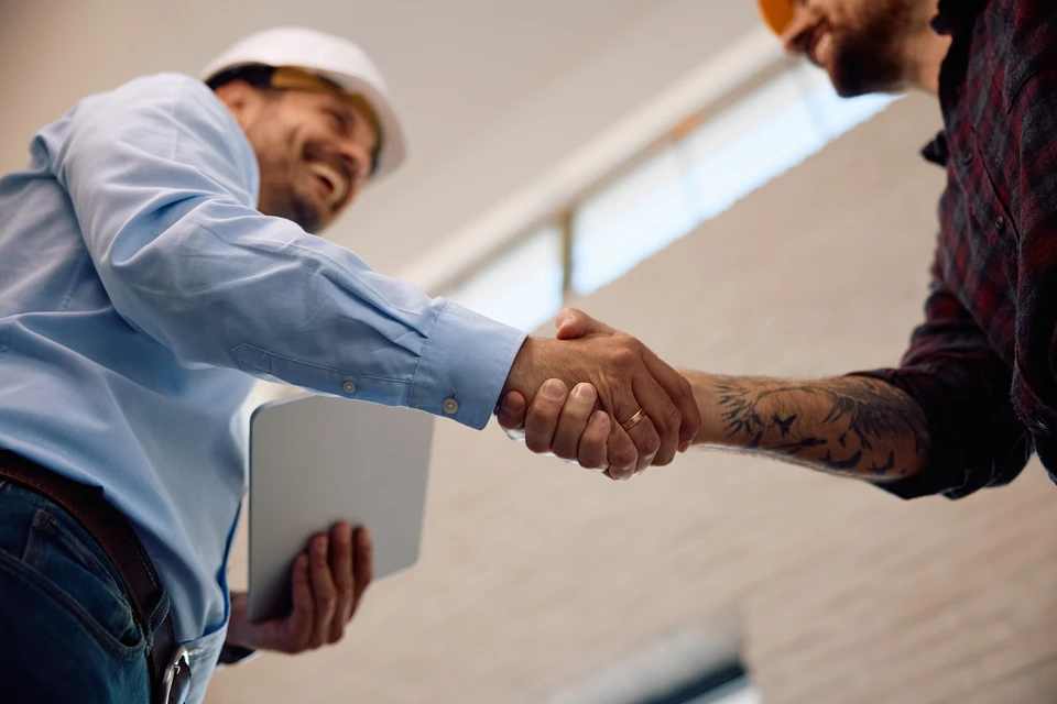 Photographie d'un professionnel menuisier-agenceur dans un bureau ou sur un chantier, en train de consulter des plans 3D sur un écran ou des documents techniques en main avec des clients ou un artisan. La scène montre une discussion collaborative autour de solutions d'aménagement, avec outils de coordination visibles (plans, relevés, maquette 3D, tableau de suivi). L'ambiance est serieuse et professionnelle, reflétant une coordination centralisée et une expertise technique au service du client.