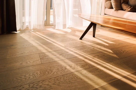 Photographie d'un artisan menuisier professionnel procédant à la pose d'un parquet massif dans un intérieur résidentiel. On voit les outils de pose (maillet, cales de bois) et une partie du parquet en cours d'installation avec un motif point de Hongrie ou pose flottante. L'artisan porte des équipements de sécurité discrets. Le contexte montre clairement un intérieur moderne en chantier ou en finition, avec des murs neutres et une lumière naturelle. L'image valorise le savoir-faire artisanal et la rigueur de la pose professionnelle, en cohérence avec le positionnement haut de gamme d'Into The Wood.