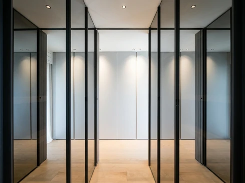 Meuble de rangement sur mesure blanc laqué intégré dans une niche murale, présentant des tiroirs à sortie totale avec système de fermeture soft-close et des poignées linéaires minimalistes. Quincaillerie Blum visible et de qualité. Derrière un tiroir partiellement ouvert, on aperçoit l'intérieur organisé avec étagères réglables et compartimentage optimisé. Éclairage LED encastré soulignant les contours. Environnement lumineux épuré, parquet clair, murs neutres. Photo prise à angle légèrement en contre-plongée pour valoriser l'aménagement modulaire et la précision de la fabrication artisanale.